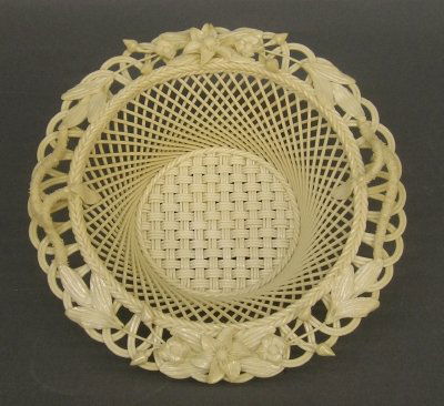 Belleek Basket: An Irish Belleek woven porcelain basket. "Clematis" pattern with an Ivory glaze with applied floral decoration. Applied cushion with Blue printed mark. Slight wear. 8 1/2" long. ESTIMATE $200-300