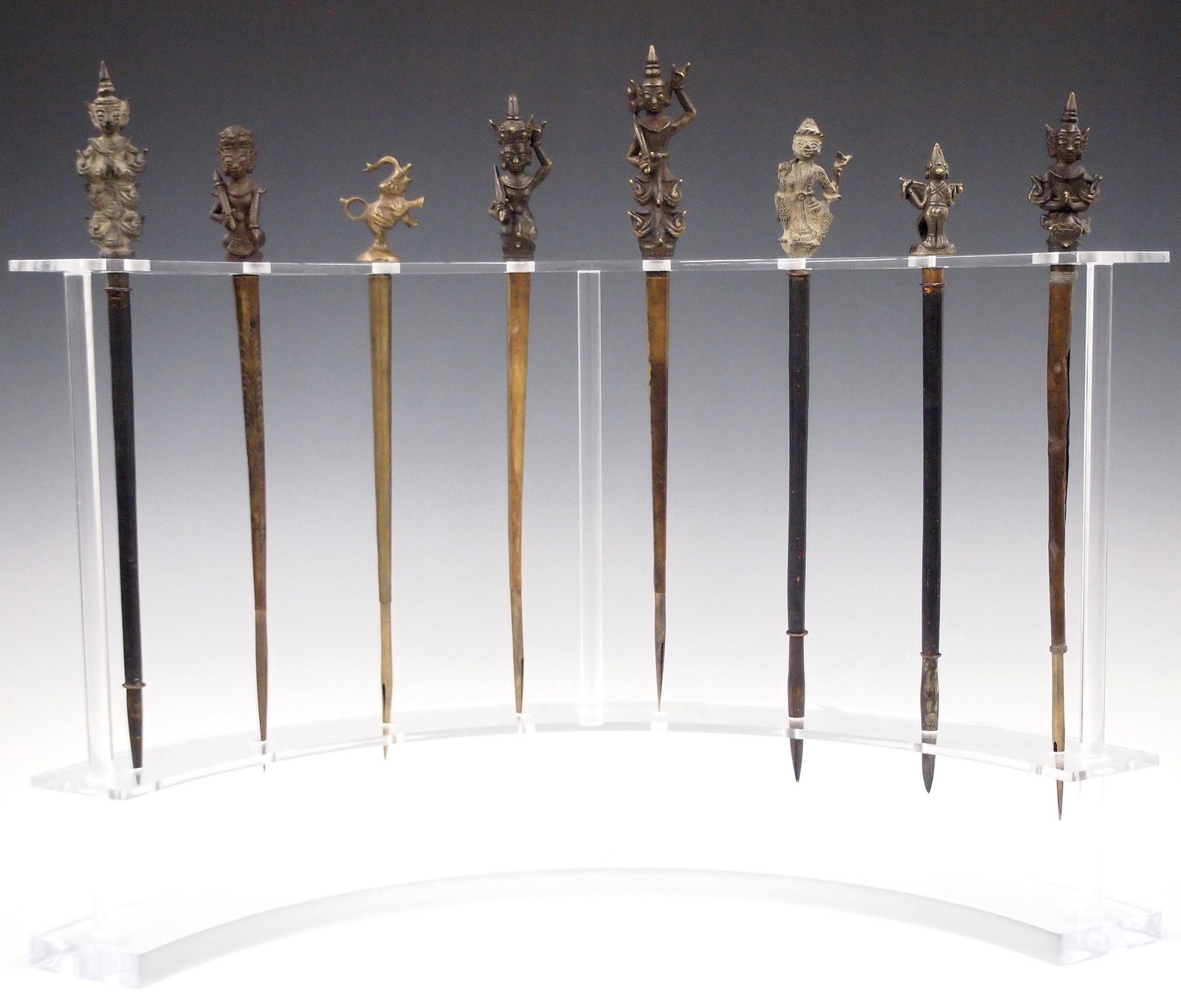 8 Burmese Tattoo Pens: Eight early 20th century Burmese tattoo pens. Hollow Brass pens with cast figural finials and a crescent shaped Lucite stand. Some surface wear, minor denting. Pens are up to 15" long, stand measures 