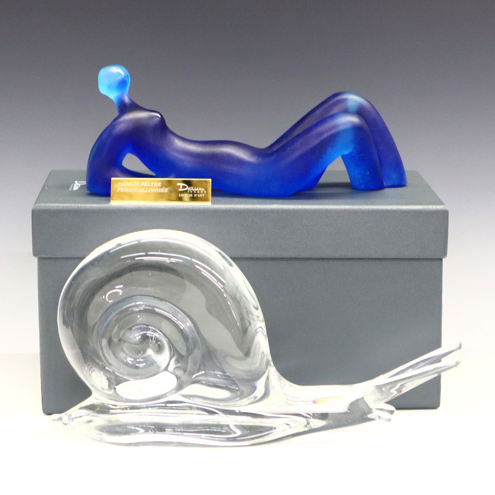 2 Daum Crystal Figures: A Daum "Femme Allongee" Pate de Verre Figure. Abstract figure of a reclining female nude in Blue, designed by Capo di Feltre. Etched "Daum France, 466/500". Minor flake on left foot. 9 1/2" long. With