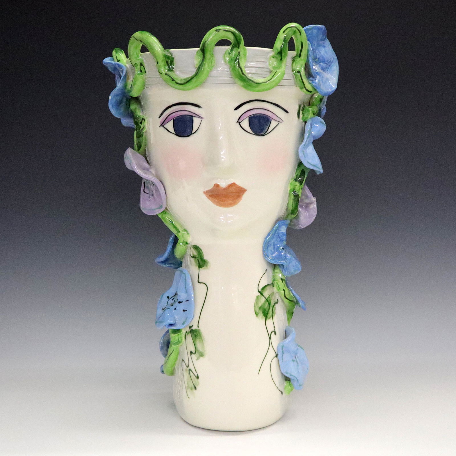 Sue Bolt Figural Vase: A late 20th century figural art pottery vase by Sue Bolt. A two-faced female bust with elongated neck, decorated in Blue and Purple floral vines. Artist signed in paint. Minor wear, small flake to edg