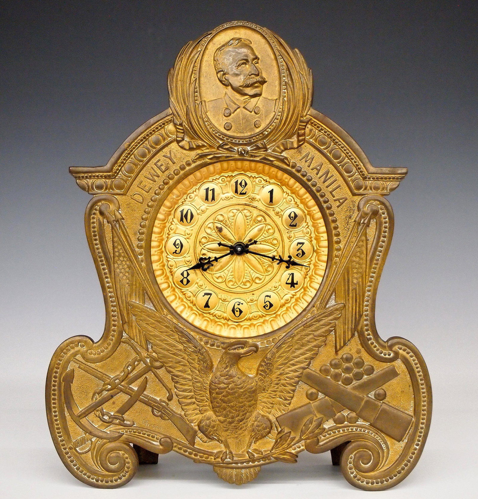 Waterbury Dewey-Manila Shelf Clock: A turn of the century Waterbury "Dewey-Manila" model shelf clock. 8-day time and strike movement, winds from the back with a Gold painted embossed dial and Black Arabic numerals. Cast Brass case marke