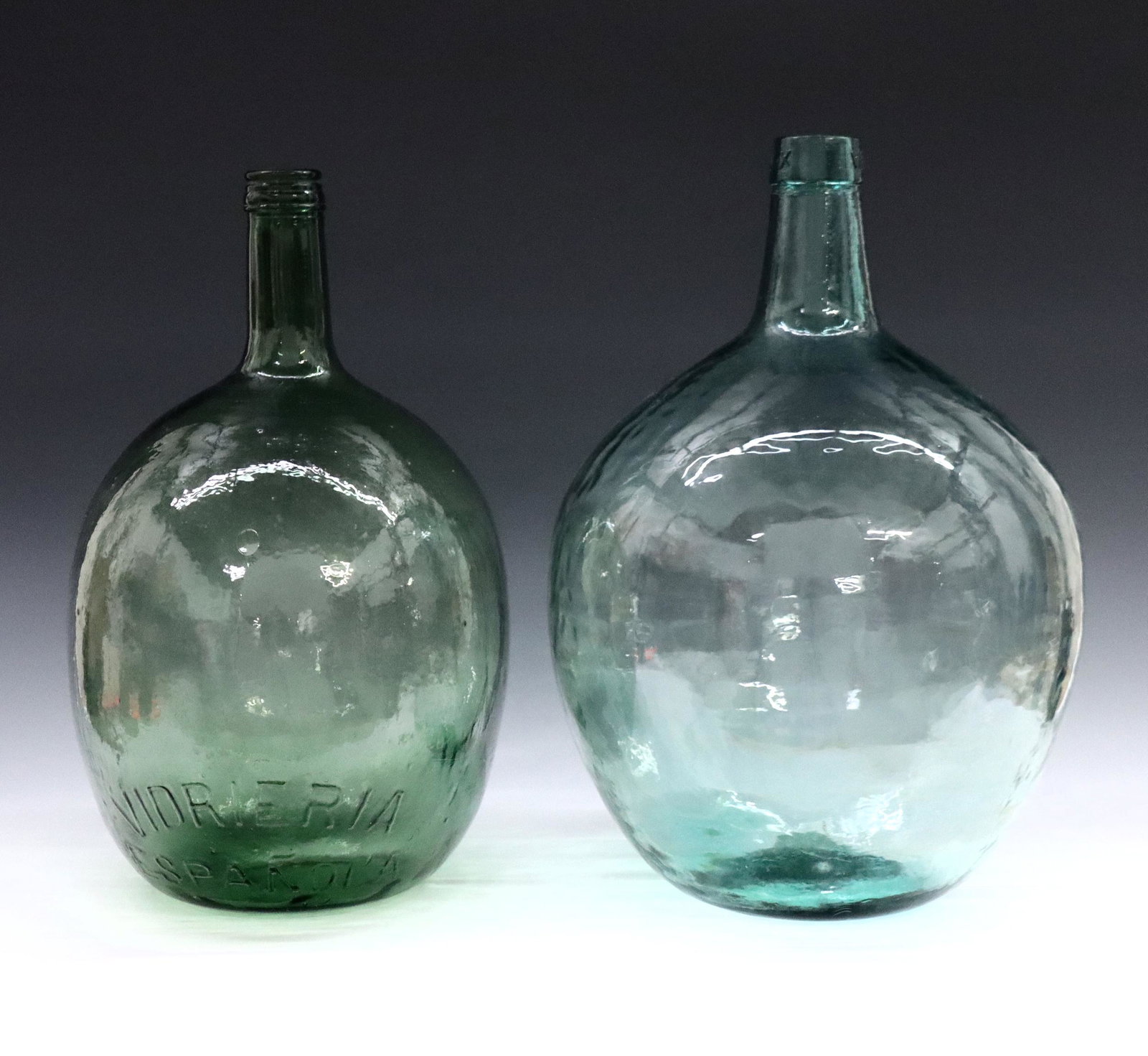 2 Spanish Demijohns: Two 20th century Spanish demijohns. Each with round forms in Green blown glass, marked "Vidrieria Espanola" and "Vieresa". Some surface wear. Up to 17" high. ESTIMATE $200-300