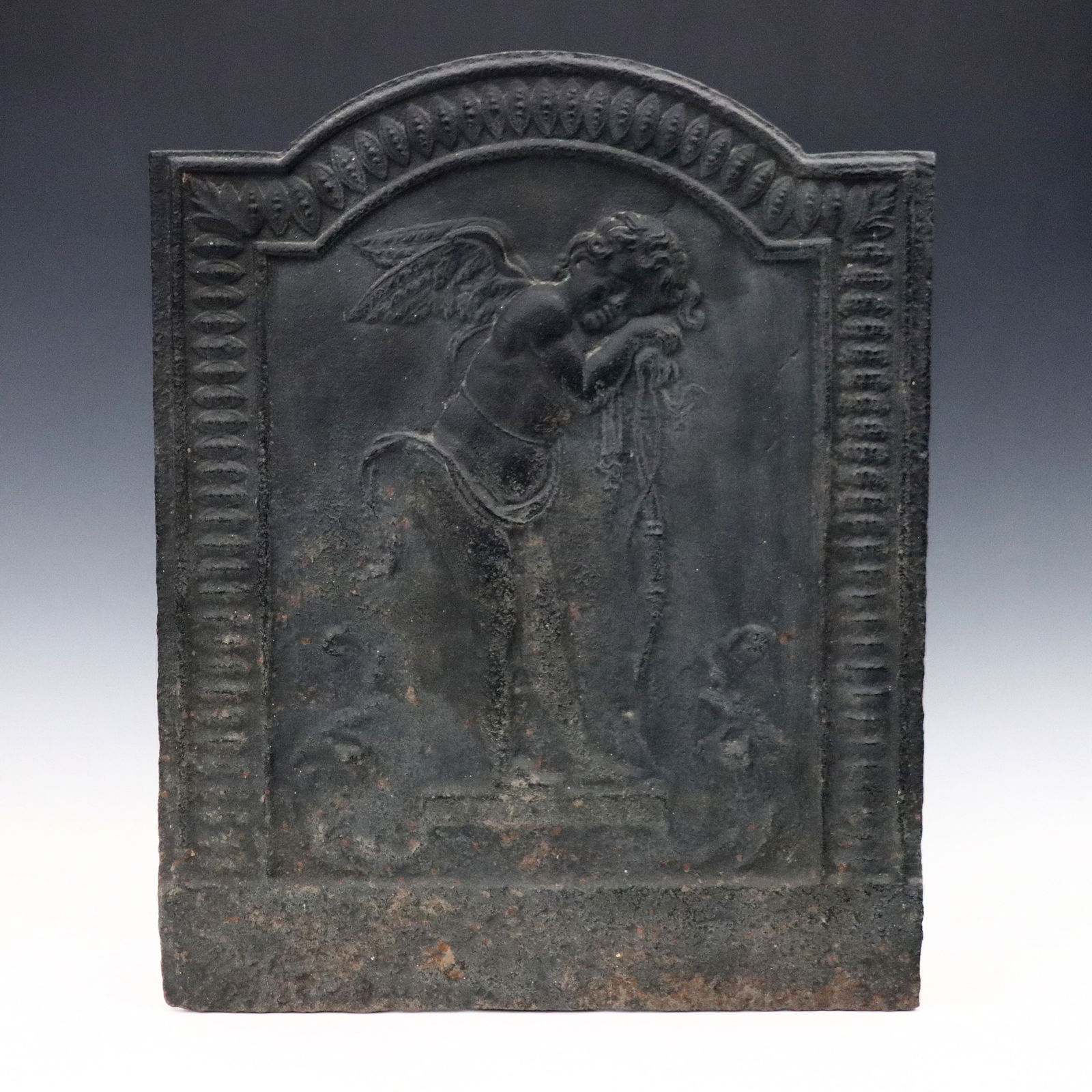 French Iron Fireback: An 18th century French cast Iron fireback. Depicts a forlorn Cupid leaning against his bow within an arched frame with floral detail. Wear, quite pitted toward bottom. 15 1/2 x 19" high overall. ESTIM