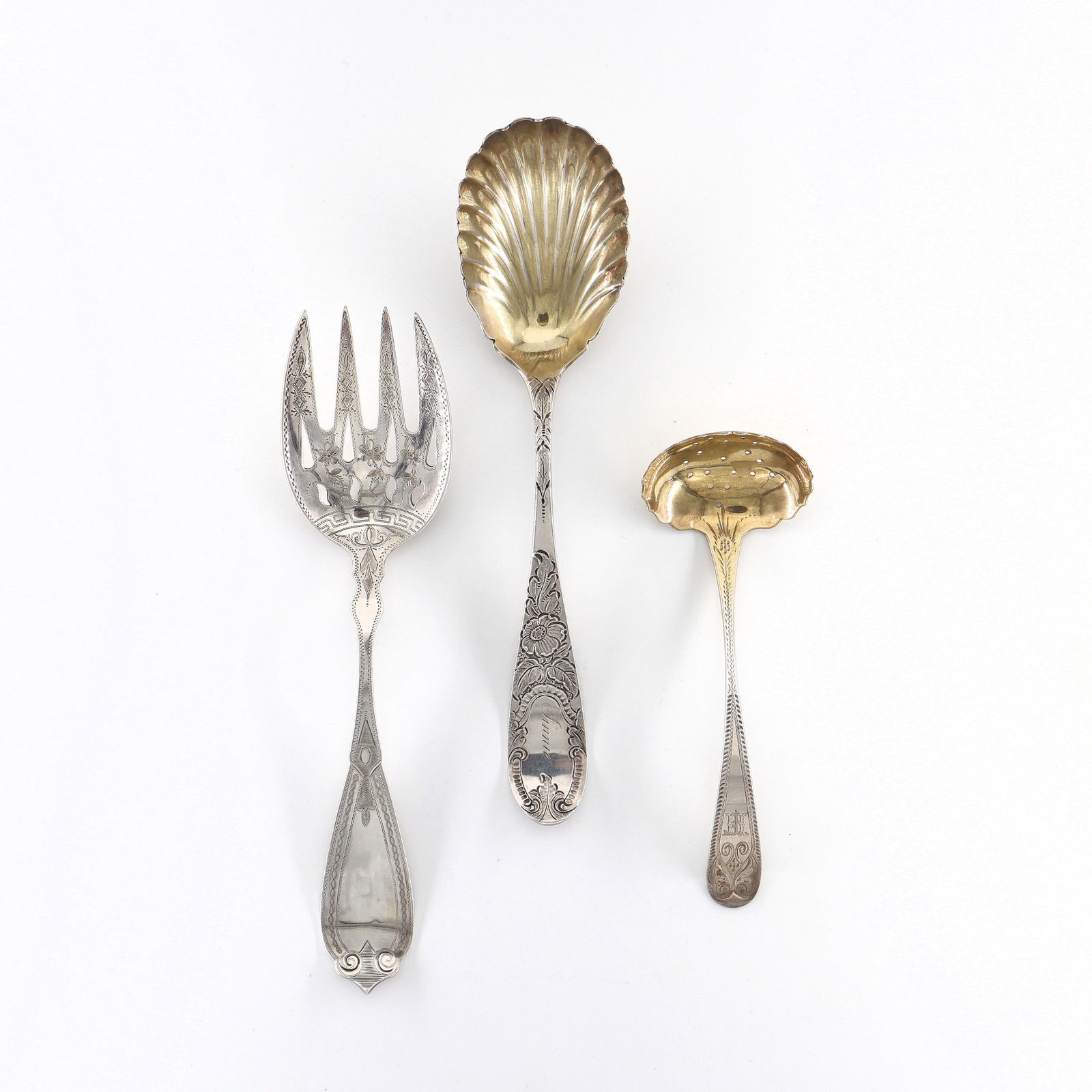 3 American Coin Silver Serving Utensils: Three 19th century American Coin Silver serving utensils with engraved decoration. Includes a pierced meat fork by Duhme, a shell form serving spoon with gold wash interior by P.B. Sadtler & Sons, and