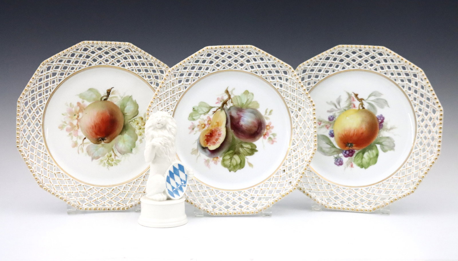 4 pc Nymphenburg Porcelain: Four pieces of 20th century Nymphenburg porcelain. Includes a seated Lion figure holding a Shield bearing the Bavarian flag, and three plates with reticulated rims and Apple design with Gilded detail.