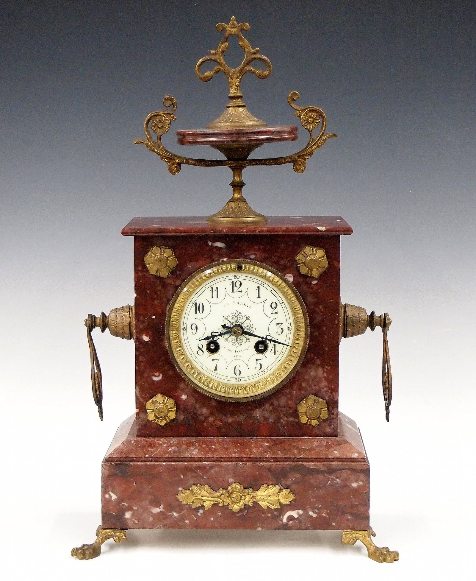French Mantel Clock: A turn of the century French mantel clock retailed by "Au Phenix, 16 Rue Lafayette, Paris". 8-day time and strike movement with convex porcelain dial and Arabic numerals. Rouge marble case with urn to