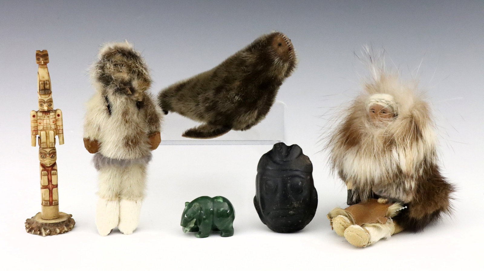 6 Inuit Items: A group of six 20th century Inuit items. Includes two dolls with fur clothing and wooden faces, a stuffed fur seal, a carved bone totem, a carved stone mask and a small carved Jade bear. Totem repaire