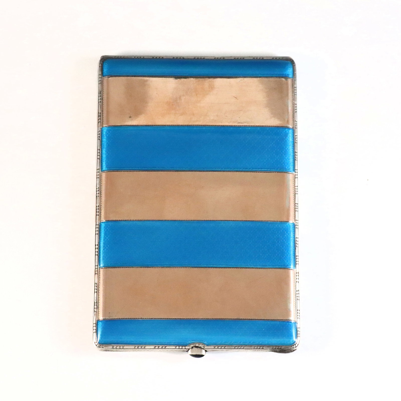 Art Deco Sterling Guilloche Cigarette Case: An early 20th century Art Deco Sterling Silver cigarette case with Guilloche enamel decoration. Striped design with Blue enamel and inset Blue glass at the clasp. Impressed "925" at interior. 6.84 tro