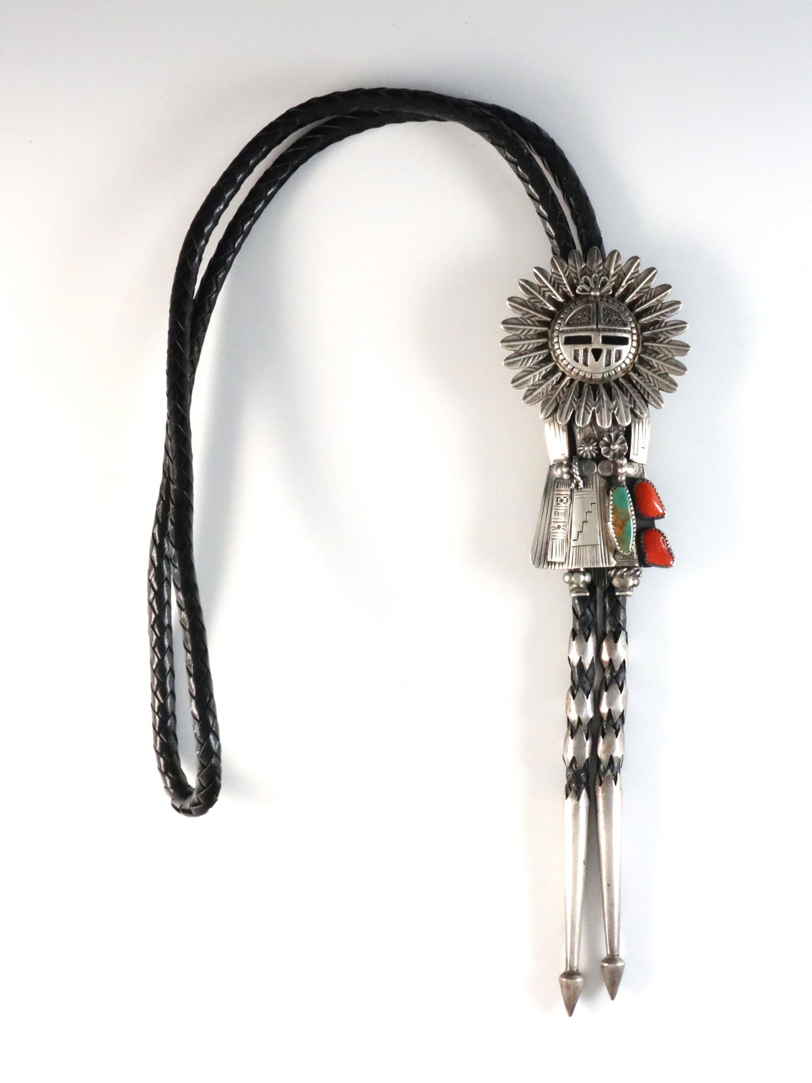 T. Byrd Navajo Sterling Bolo Tie: A vintage Navajo Sterling Silver bolo tie by Thomas Byrd. Features a figural Sun Face Kachina design with bezel set Turquoise and Coral on a braided leather tie. Marked "Sterling / Thomas Byrd / Red M