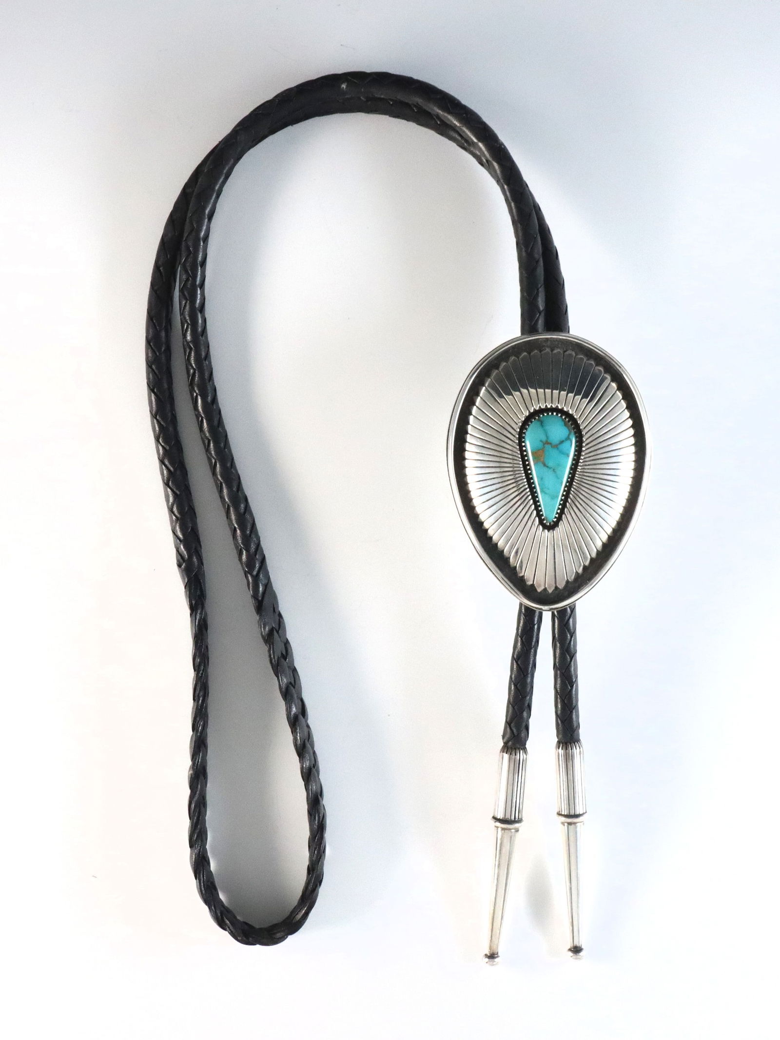 B. McCurtain Kiowa Sterling Bolo Tie: A vintage Kiowa Sterling Silver bolo tie by Byron McCurtain. Features an inverted teardrop form clasp with a sawtooth bezel set Turquoise stone and reeded design on a braided leather tie. Marked "Ster