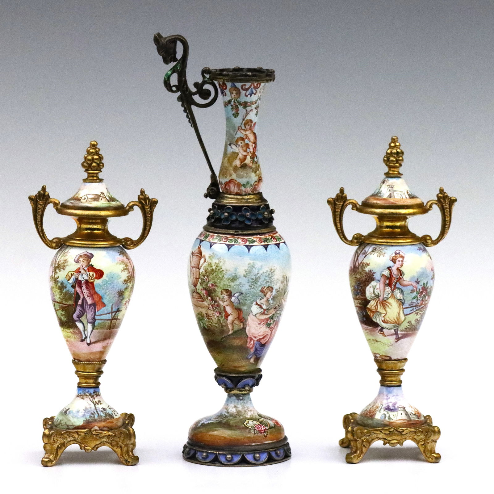 3 Viennese Miniature Enameled Vases: Three turn of the century Viennese miniature enameled vases. Hand-enameled scenic decoration depicting ladies, gentleman and cherubs on Gilded metal mounts. Includes a pair of covered urns and a balus