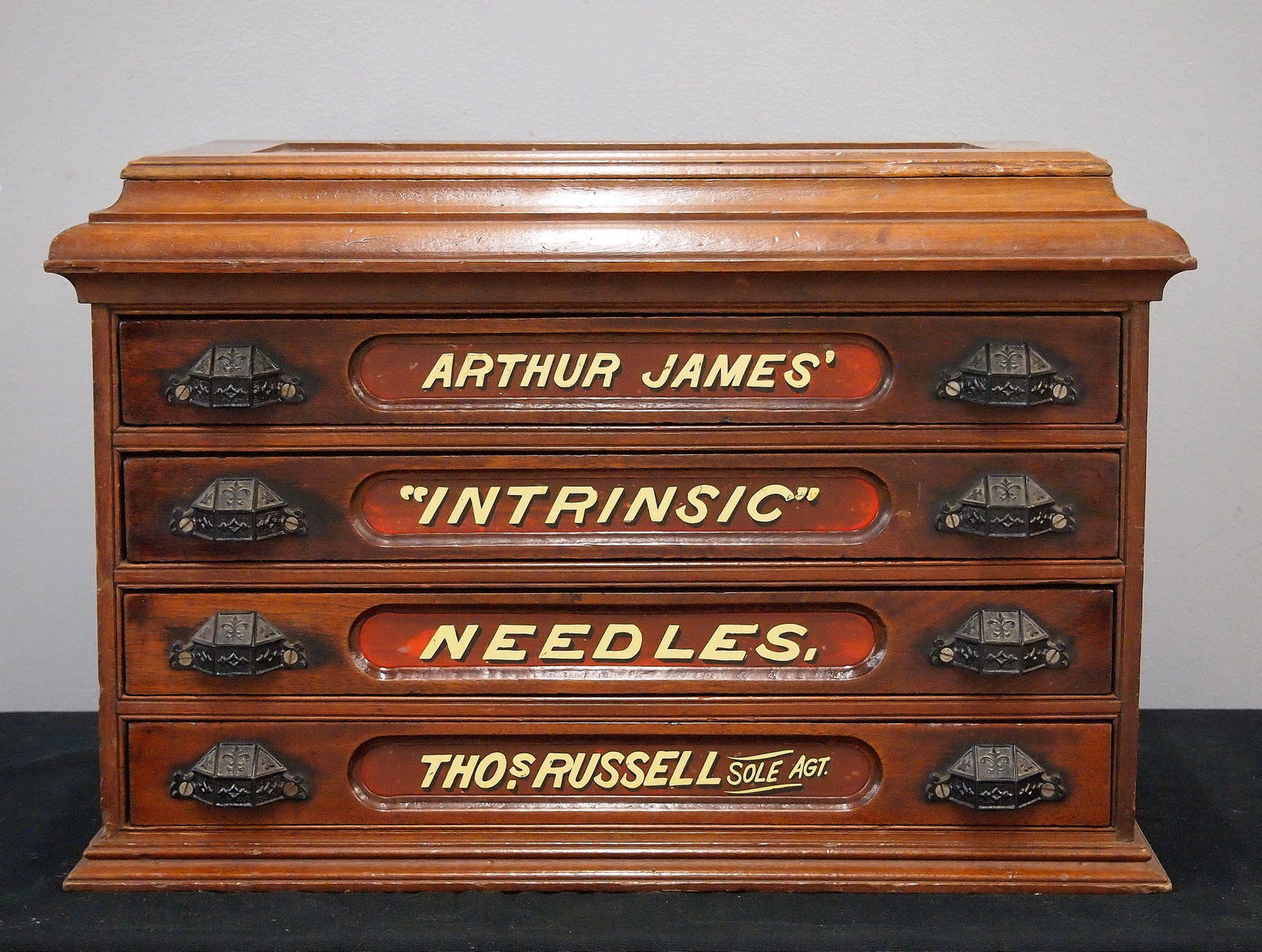 Arthur James Thread Cabinet: A turn of the century Arthur James thread cabinet. Walnut construction features a molded top with lift-up panel over four drawers with reverse painted glass labels "Arthur James, Intrinsic, Needles,