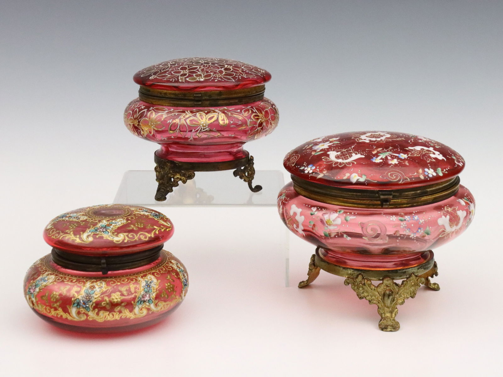 3 Bohemian Glass Dresser Boxes: Three turn of the century Bohemian Glass dresser boxes. Includes three circular Cranberry Glass boxes with Gilded metal mounts and floral enameled decoration. Each unmarked, smallest with paper label