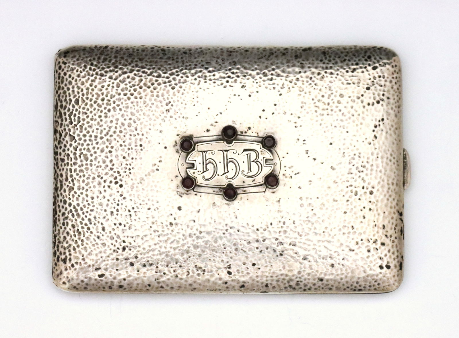 American Sterling Cigarette Case: A turn of the century American Sterling Silver cigarette case by James E. Blake Co. Hinged rectangular form with hammered exterior and monogrammed plaque surrounded by six faceted Red jewels. Marked "