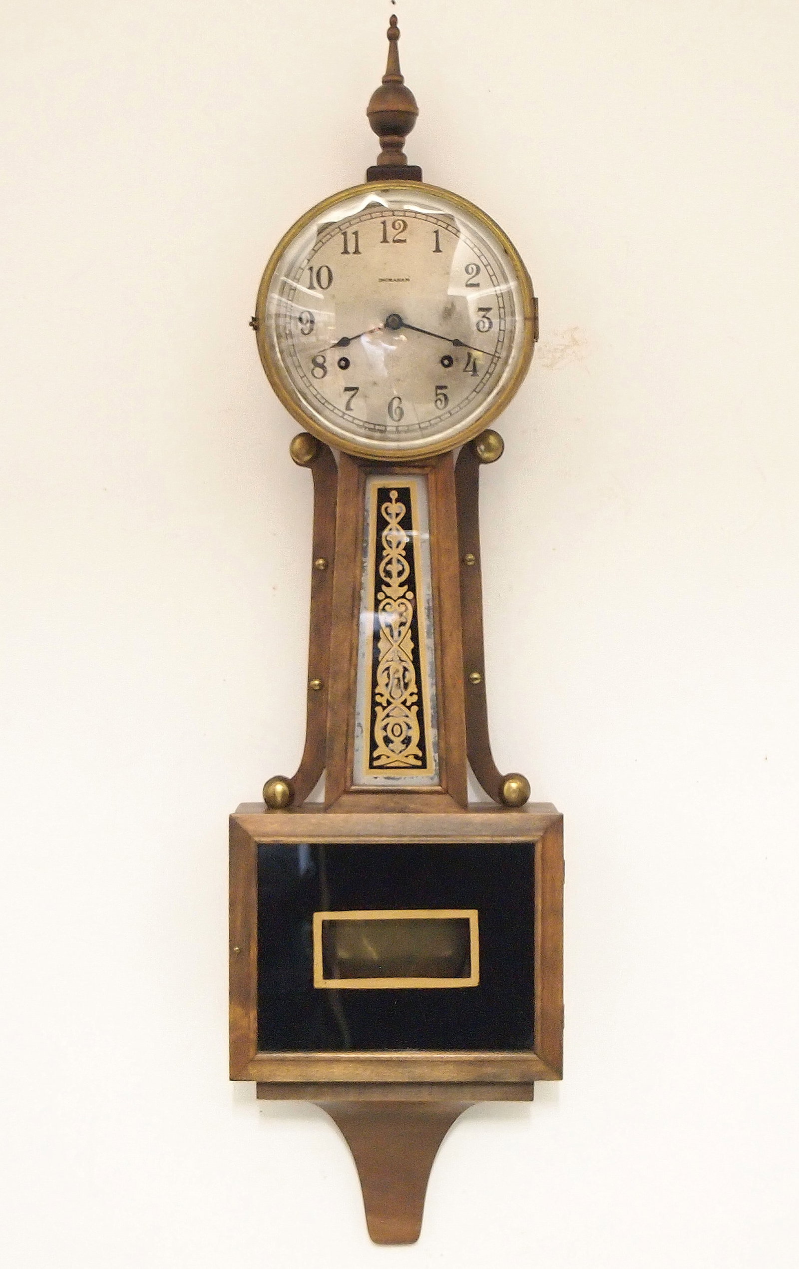 Ingraham Banjo Clock: A 1930's Ingraham Banjo clock. 8-day time and strike movement with Silvered dial and Arabic numerals. Birch case with turned finial, round top, shaped arms and reverse painted throat and lower door
