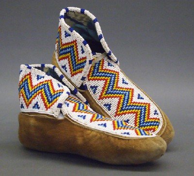 Pr. Cree Moccasins: A Pair of 20th Century Cree Moccasins. With beaded on vamps and cuffs having a a zigzag pattern in red, yellow, and two shades of blue against a white background. Some wear, both with small holes at t