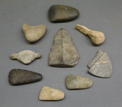 9 Stone Implements: A Group of Nine Native American Stone Implements. Includes spear heads, axe heads, and other implements. Wear to each, one (in the form of a bird's head) has an old repair. From 2" to 5" long. ESTIMAT