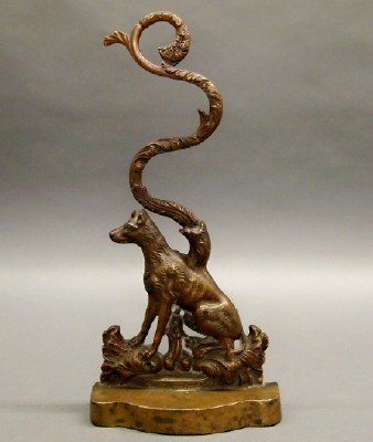 Dog Form Doorstop: A 19th Century Bronze Clad Cast Iron Doorstop. Single figure of a dog, shown seated, with scrolling leaf design protruding from its mid-back, resting on a serpentine base. Unmarked. Minor wear. 13 3/4